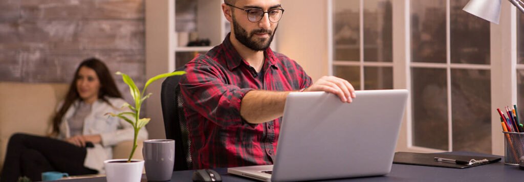 Bearded freelancer opening his laptop to start working on living room. girlfriend relaxing on sofa watching tv in the background - Apply for Dubai Freelance Visa now